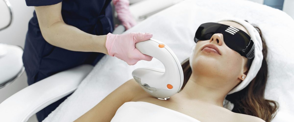 Crop faceless woman in medical uniform and gloves using laser tool on young woman wearing safety glasses and towel lying on couch in hospital